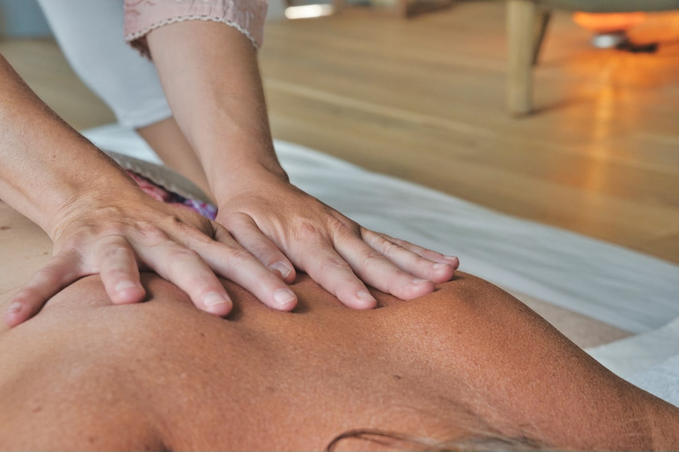 A person receives a back massage while lying face down on a white surface. Another person's hands are gently pressing on their upper back in a calm, sunlit room with wooden floors.