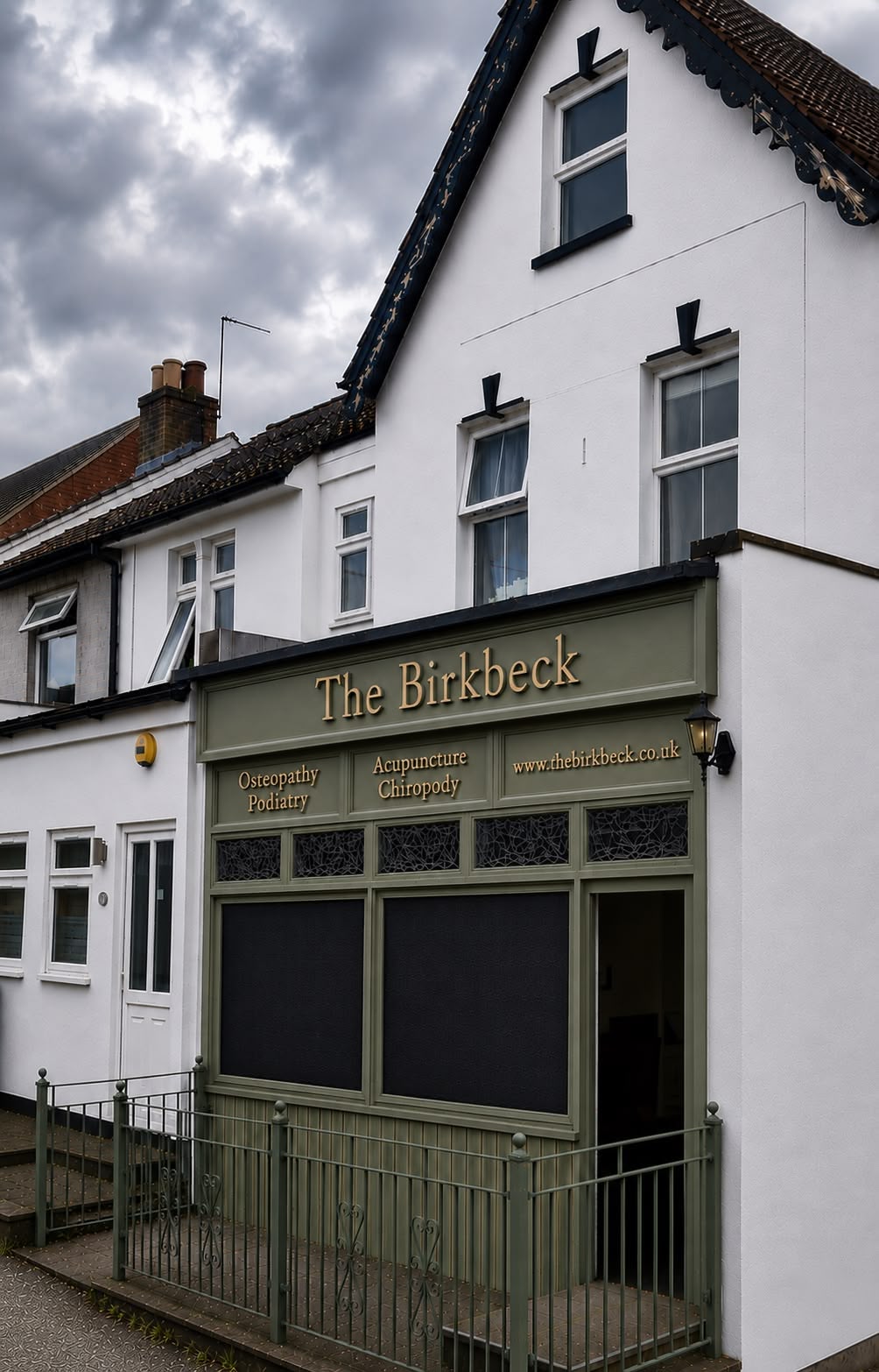 A white building with a green shopfront labeled The Birkbeck, offering osteopathy, podiatry, acupuncture, and chiropody services. The windows above the shop are white-framed, and the sky is cloudy.