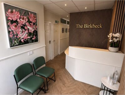 A modern waiting room with green chairs, a white curved reception desk, a wall sign reading The Birkbeck, a pink floral painting, wood panelling, potted orchids, and a small table with water and glasses.