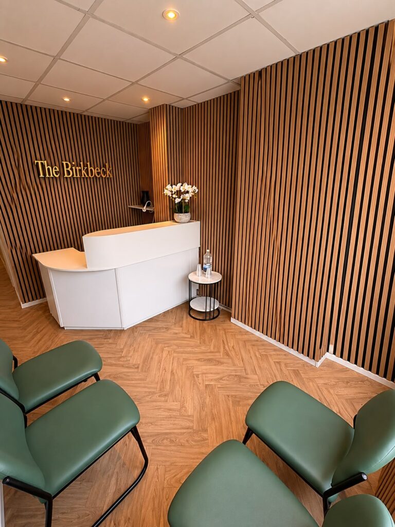 A modern waiting room with green chairs, a white curved reception desk, a round side table with water, and vertical wooden slat walls. A sign reads The Birkbeck and a vase of white flowers is on the desk.