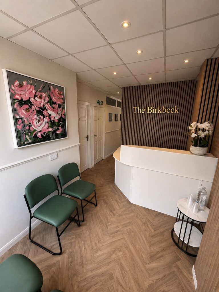 A modern waiting room with green chairs, a white reception desk labelled The Birkbeck, a floral painting, wood accents, potted orchid, and water glasses on a small round table.
