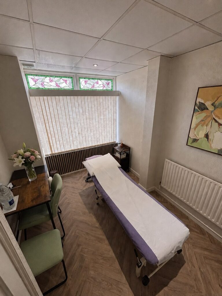 A treatment room with a reclining examination couch covered in white and purple paper, two green chairs, a wooden desk with flowers, and a painting on the wall. Vertical blinds cover windows with stained glass panels above.