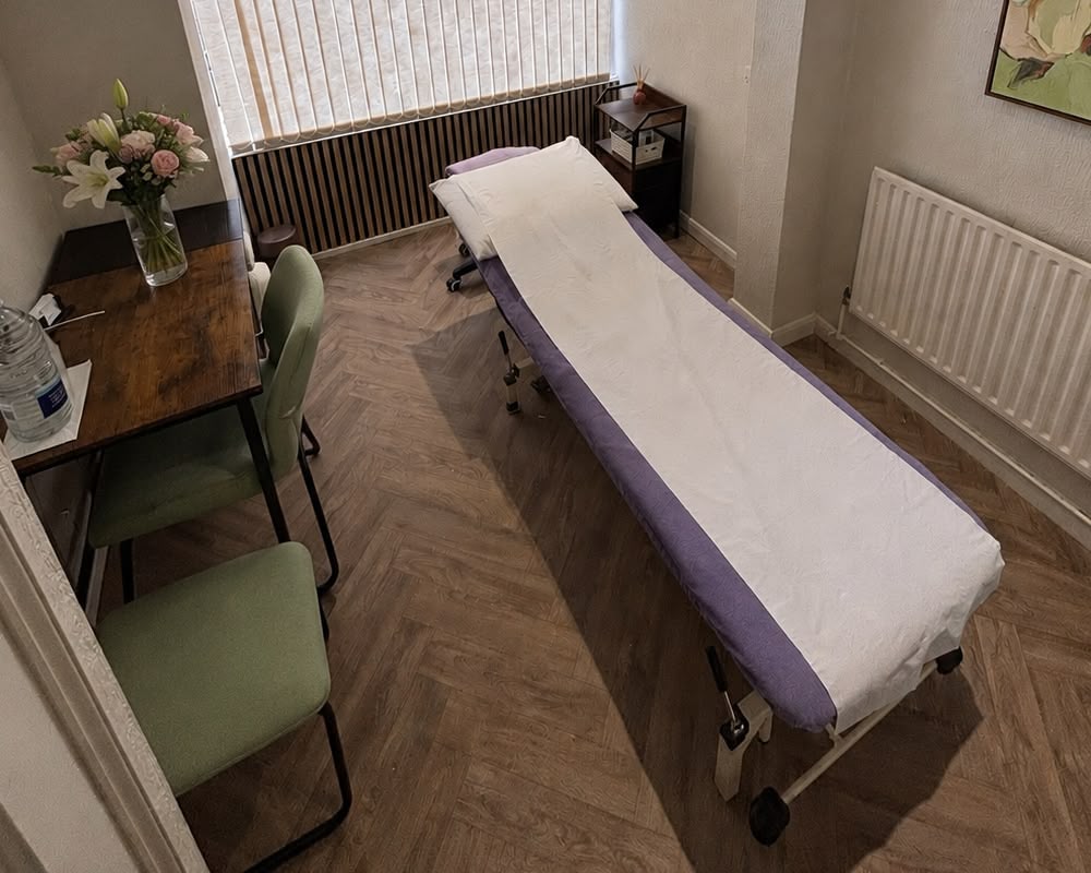A massage therapy room with a treatment table covered in white and purple sheets, two green chairs by a wooden desk with flowers, a water bottle, and a window with vertical blinds letting in natural light.